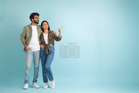 Photo for Loving cheerful young eastern couple wearing casual outfits looking and pointing at copy space for advertisement on blue colored studio background, full length. Great offer for two - Royalty Free Image