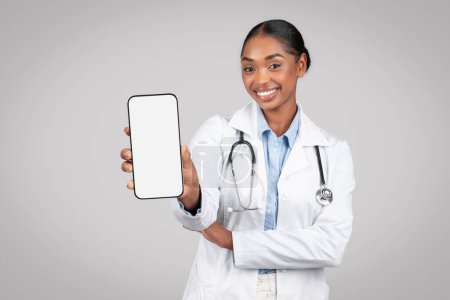Photo for Cheerful black woman doctor in white coat hold smartphone with empty screen, isolated on gray background studio, expressing positivity. Healthcare services, ad and offer, app recommendation - Royalty Free Image