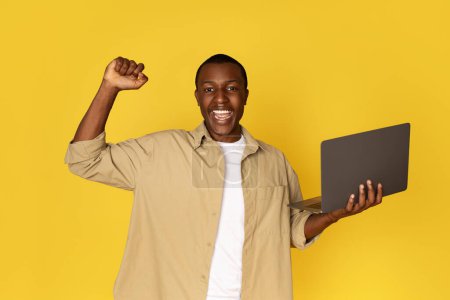 Photo for Glad excited millennial african american guy in casual with laptop celebrating victory, rejoices success, isolated on yellow studio background. Winner with technology, work and study remotely - Royalty Free Image
