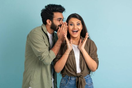Photo for Handsome young middle eastern guy sharing secret or whispering gossips into his girlfriends ear, excited pretty brunette indian woman gesturing, isolated on blue studio background, copy space - Royalty Free Image