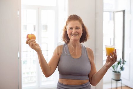 Téléchargez les photos : Concept de nutrition saine. Femme âgée sportive souriante tenant la pomme et le verre avec du jus, dame âgée heureuse en vêtements de sport recommandant une boisson vitaminée pour un régime alimentaire sain, bénéficiant du bien-être - en image libre de droit