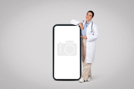 Photo for Latin woman doctor in uniform, standing next to huge cellphone with blank screen, allowing for custom message insertion, against grey studio background, symbolizing telehealth - Royalty Free Image