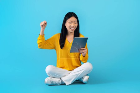 Foto de Alegre joven mujer china feliz sentado en el suelo, jugando videojuegos en la tableta digital, celebrando el éxito de la victoria, mirando la pantalla de la almohadilla, levantando la mano y exclamando, aislado en el fondo azul - Imagen libre de derechos