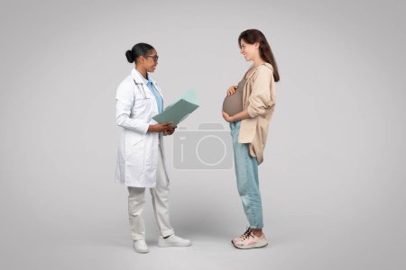 Photo for Glad black woman doctor consults pregnant patient, discussing healthcare, nutrition and wellbeing during pregnancy, isolated on gray background studio. Support in clinic, medicine - Royalty Free Image