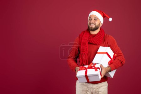Téléchargez les photos : Souriant homme caucasien millénaire avec barbe dans le chapeau Santa tenir des boîtes de cadeaux, regarder l'espace de copie, isolé sur fond studio bordeaux. Noël, Nouvel An vente, idée cadeau - en image libre de droit