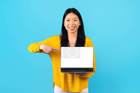 Photo for Online entertainment concept. Happy young asian woman digital nomad showing laptop computer with white empty screen, blue background, mockup, copy space. E-education, online course - Royalty Free Image