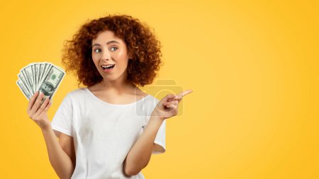 Photo for Smiling funny european teen lady, hold money, point finger at free space, isolated on yellow background. Recommendation financial profit and win, credit advice, ad and offer - Royalty Free Image