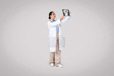 Photo for Focused latin woman doctor examining X-ray of the lungs, reflecting a moment of medical evaluation and diagnosis, posing against grey studio background - Royalty Free Image
