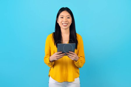Foto de Positivo alegre mujer asiática joven utilizando tableta digital sobre fondo de estudio azul, sonriendo a la cámara, compras, chat en línea. Feliz señora china que aprende el idioma extranjero en línea, e-educación - Imagen libre de derechos