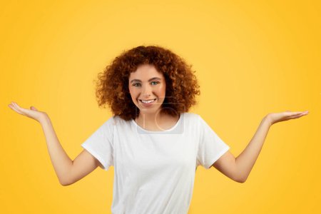 Photo for Cheerful pretty teen woman in white t-shirt hold on hands free space, isolated on yellow background, studio. Student enjoy youthful, choose, lifestyle, ad and offer, question - Royalty Free Image