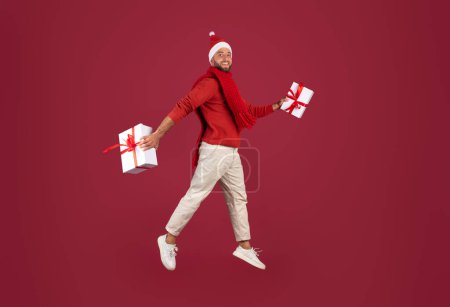 Foto de Glad millennial hombre caucásico en Santa sombrero salta, congelado en el aire con cajas de regalos, se divierte, aislado en el fondo del estudio de color burdeos. Feliz Navidad, venta de Año Nuevo, idea de regalo, anuncio y oferta - Imagen libre de derechos