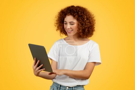 Téléchargez les photos : Glad teen femme européenne utilise tablette, naviguant à travers les pages numériques pour l'étude et le travail, isolé sur fond jaune. Absorbé dans les tâches, incarnent l'intersection de la jeunesse, la technologie, la productivité - en image libre de droit