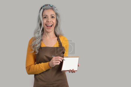Photo for Surprised glad senior woman gray hair framer in apron hold banner with empty space, isolated on gray studio background. Farming, healthy lifestyle, ad and offer, agro eco business - Royalty Free Image