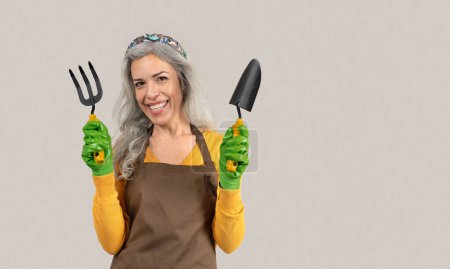 Photo for Engaged in garden, senior woman with gray hair tends to vegetables, flowers with shovel in hand, isolated on gray studio background. Lifestyle, hobby, active retirement at free time - Royalty Free Image