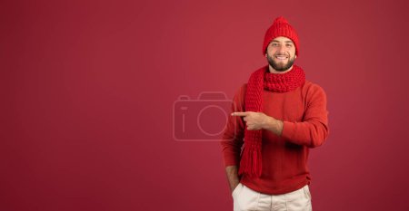 Foto de Feliz chico caucásico milenario guapo con barba en el dedo del sombrero en el espacio libre, aislado en el fondo del estudio de color burdeos, panorama. Recomendación para la venta, el anuncio y la oferta de invierno - Imagen libre de derechos