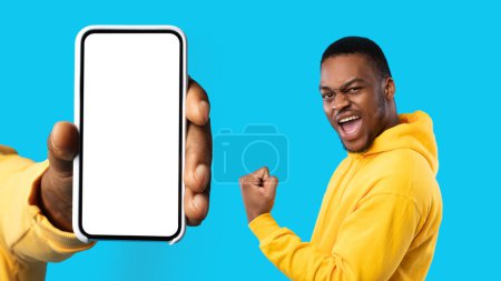 Photo for Happy excited young black guy show first, celebrate win, success, near hand with phone with empty screen, isolated on blue background, studio. Good news emotions, business, work app - Royalty Free Image