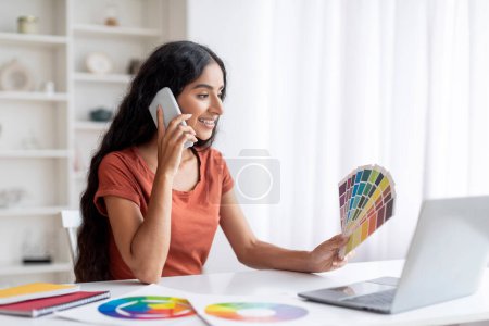 Photo for Engaged creative millennial indian woman freelancer, calmly managing her design projects while balancing between laptop use and phone conversations at home office, holding color palette - Royalty Free Image
