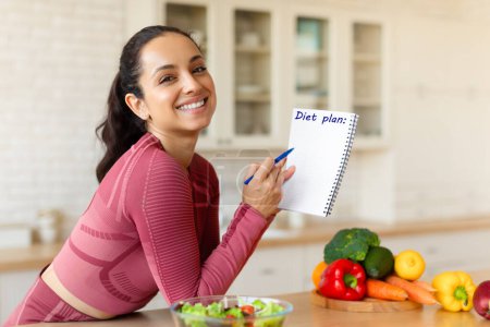 Fit lächelnde Frau posiert mit Diätplan, schreibt Menüs und gesunde Abnehmrezepte in Notizbuch, steht in der modernen Küche drinnen, schaut in die Kamera. Schlankheitskonzept