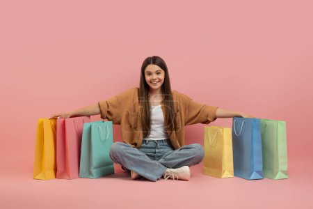 Photo for Happy Teen Girl Surrounded By Bright Shopping Bags Sitting Over Pink Studio Background, Smiling Cute Female Teenager Enjoying Fashion Sales, Embracing Shopper Bags And Looking At Camera, Copy Space - Royalty Free Image