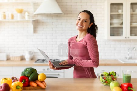 Photo for Fit lady using laptop planning healthy meals standing in modern kitchen, researching and planning nutritious recipes for weight loss. Healthy nutrition and wellbeing concept - Royalty Free Image