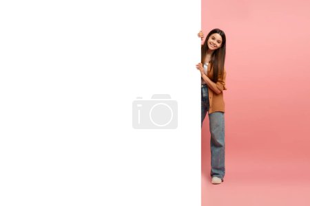 Foto de Interesante oferta. Chica adolescente feliz asomándose detrás del tablero de publicidad blanco para texto o diseño, sonriente adolescente sosteniendo la cartelera, mirando a la cámara, de pie sobre el fondo rosa - Imagen libre de derechos