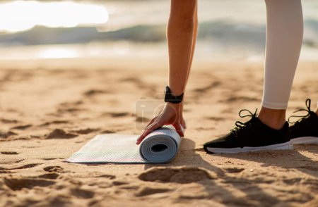 Photo for Fit lady putting mat on sand, preparing for training on ocean beach at spare time on sunny day, closeup, side view. Fitness, body care, weight loss and sport training outdoors - Royalty Free Image