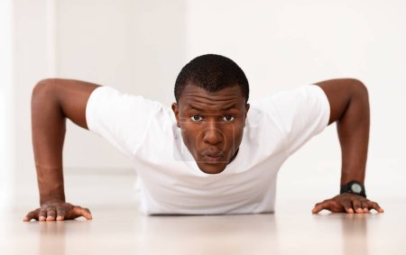 Téléchargez les photos : Motivé athlétique millénariste noir gars travaillant sur les bras et la force des muscles de la poitrine, faire des pompes, regarder la caméra. Sportif afro-américain faisant de l'exercice à la maison, profitant de l'entraînement - en image libre de droit