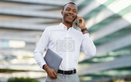 Téléchargez les photos : Appel d'affaires. Homme d'affaires noir souriant parlant sur un téléphone portable tout en marchant à l'extérieur près du centre de bureau moderne, bel entrepreneur afro-américain tenant un ordinateur portable, bénéficiant d'une conversation agréable - en image libre de droit