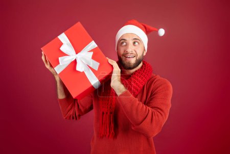 Téléchargez les photos : Curieusement heureux drôle mec caucasien millénaire avec barbe en chapeau Santa écouter cadeau boîte, profiter des vacances, isolé sur fond de studio bordeaux. Présent à Noël, Nouvel An - en image libre de droit