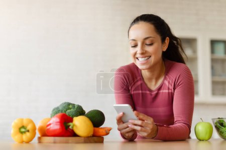 Photo for Slimming App. Young Fit Lady In Sportswear Using Weight Loss Application On Cellphone Cooking Healthy Meals In Modern Kitchen Indoors, Posing With Smartphone And Websurfing - Royalty Free Image