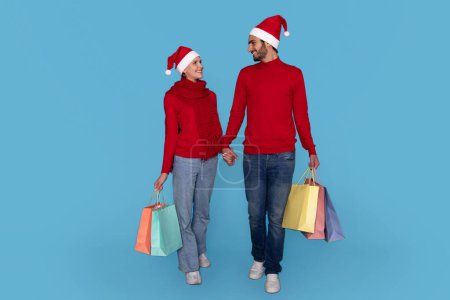 Photo for Holiday Sales. Happy Young Couple Wearing Santa Hats Walking With Shopping Bags Over Blue Studio Background, Cheerful Man And Woman Enjoying Seasonal Offers And Discounts, Copy Space - Royalty Free Image
