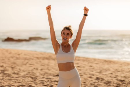 Foto de Mujer atleta europea satisfecha en ropa deportiva levantando las manos, celebra gran resultado de entrenamiento, el éxito en la playa del mar. Deporte al aire libre, acabado, emociones ganadoras - Imagen libre de derechos