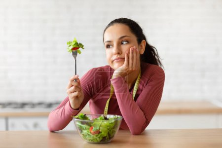 Téléchargez les photos : Diet Struggles. Femme mince insatisfaite en vêtements de fitness regardant fourchette ayant salade de légumes pour le dîner, posant fatigué de suivre un régime et une alimentation saine dans la cuisine moderne à la maison. Menu de perte de poids - en image libre de droit
