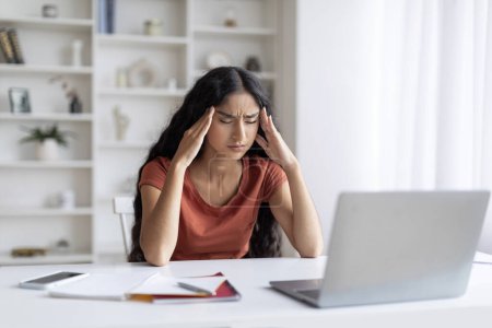 Photo for Exhausted young indian lady sitting at workplace in front of laptop, eyes closed, touching her head, suffer from migraine, surrounded by scattered papers, long hours spent on her home office tasks - Royalty Free Image