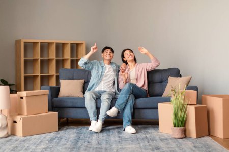 Photo for Asian couple surrounded by moving boxes discussing plans for upcoming home renovations, sitting on sofa gesturing dreaming about nice interior amidst their packed belongings indoor - Royalty Free Image