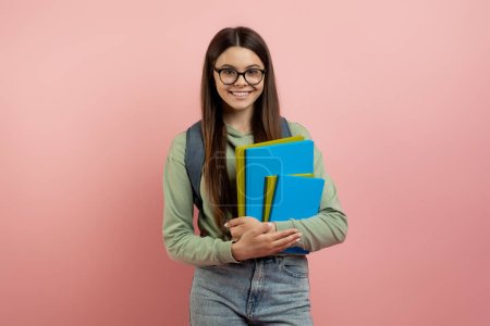 Foto de Retrato de estudiante adolescente sonriente con mochila y libros de trabajo posando sobre fondo rosa en el estudio, adolescente linda feliz en gafas mirando a la cámara, listo para la escuela, disfrutando del estudio - Imagen libre de derechos