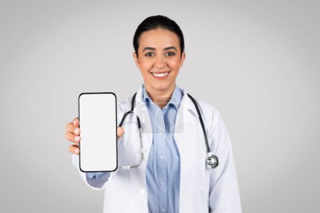 Photo for Happy latin woman doctor presents cellphone with blank screen towards the camera, seamlessly integrating space for medical offer or promotional message, grey studio background - Royalty Free Image