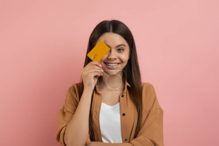 Photo for Happy teenage girl holding credit card, covering eye and smiling at camera, cheerful beautiful teenager female recommending trustful bank, standing over pink background in studio, copy space - Royalty Free Image