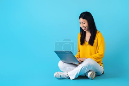 Téléchargez les photos : Sourire positif jolie jeune femme asiatique entrepreneur indépendant travaillant sur ordinateur portable, assis sur le sol sur fond bleu studio. Dame chinoise étudiant en ligne, en utilisant l'ordinateur, l'espace de copie - en image libre de droit