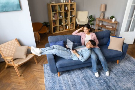 Téléchargez les photos : Les conjoints japonais aiment regarder une vidéo en ligne sur leur ordinateur portable, se détendre sur le canapé dans le confort du salon à la maison. Technologie en ligne et loisirs en informatique familiale - en image libre de droit