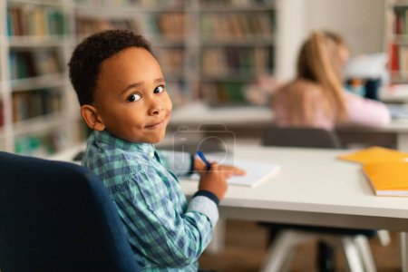 Téléchargez les photos : Amical écolier afro-américain, assis à son bureau de classe, tourne le dos, regardant et souriant à la caméra, montrant un moment de joie et d'ouverture pendant son éducation primaire - en image libre de droit