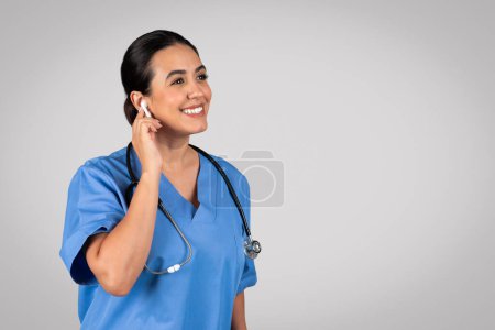 Téléchargez les photos : Femme latine médecin avec oreillette dans l'oreille, s'engage dans une consultation en ligne avec le patient, incarnant le professionnalisme de la télésanté et la communication numérique, sur fond de studio gris, espace de copie - en image libre de droit