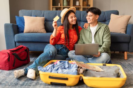 Téléchargez les photos : Joyeux couple coréen en utilisant un ordinateur portable réservation de vacances en ligne, le paiement des billets et de l'hôtel avec carte de crédit à l'intérieur de la maison confortable. Préparation des voyages et concept de commerce électronique - en image libre de droit