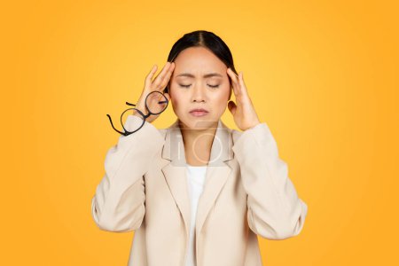 Photo for Asian woman, adorned in professional suit, endures moment of pain from migraine while at her work environment. Her expression reflects struggle of maintaining professionalism amidst discomfort - Royalty Free Image