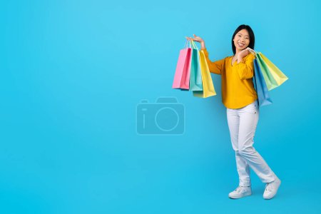 Photo for Happy pretty young korean woman wearing casual outfit showing her purchases colorful shopping bags, isolated on blue studio background, full length, copy space for advertisement. Retail, consumerism - Royalty Free Image