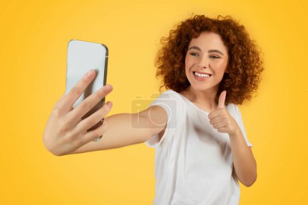 Téléchargez les photos : Femme adolescente souriante prend avec enthousiasme selfie au téléphone, faire signe doigt vers le haut, reflétant moment de joie et d'affirmation. Style auto-photographie sur fond jaune, studio - en image libre de droit