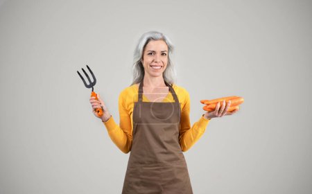 Photo for Smiling senior woman farmer in apron hold garden rake, carrots, enjoy eco harvest, isolated on gray background. Healthy food, agro business, farmer work, ad and offer, hobby - Royalty Free Image