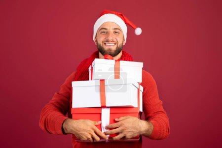 Téléchargez les photos : Joyeux beau millénium homme caucasien dans le chapeau Santa tenir des boîtes de cadeaux, profiter des vacances, Noël, la vente du Nouvel An, isolé sur fond de studio bordeaux. Présente pour un événement de fête familiale - en image libre de droit