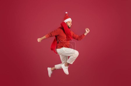 Foto de Positivo hombre caucásico milenario en Santa sombrero salta, congelado en el aire, disfrutar de las vacaciones, se divierte, aislado en el fondo del estudio de color burdeos. Navidad, Año Nuevo venta emociones, estilo de vida - Imagen libre de derechos