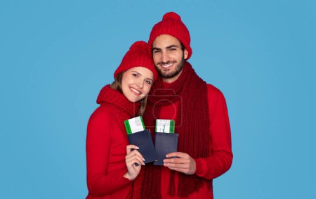Foto de Viajes de invierno. Feliz pareja joven sosteniendo pasaportes con boletos de tarjeta de embarque, hombre alegre y mujer en sombreros de punto sonriendo a la cámara, posando aislado en el fondo del estudio azul, espacio de copia - Imagen libre de derechos